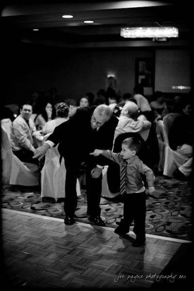 raleigh wedding photographer - jenna and ryan A black and white photo of an older man and a young boy dancing on a wooden floor. The boy, dressed in a shirt and tie, extends his arm toward the man, who is bending slightly, guiding him. In the background, people are seated at tables.