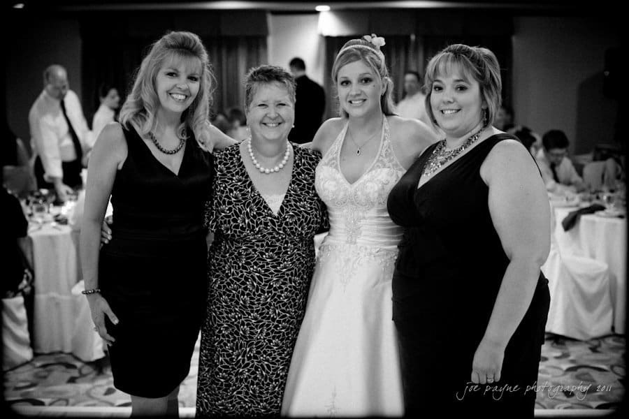 raleigh wedding photographer - jenna and ryan Four women are posing together at a formal event. The woman in the center wears a wedding dress, while the others are in elegant black dresses. They are smiling, and a crowded reception hall is visible in the background.