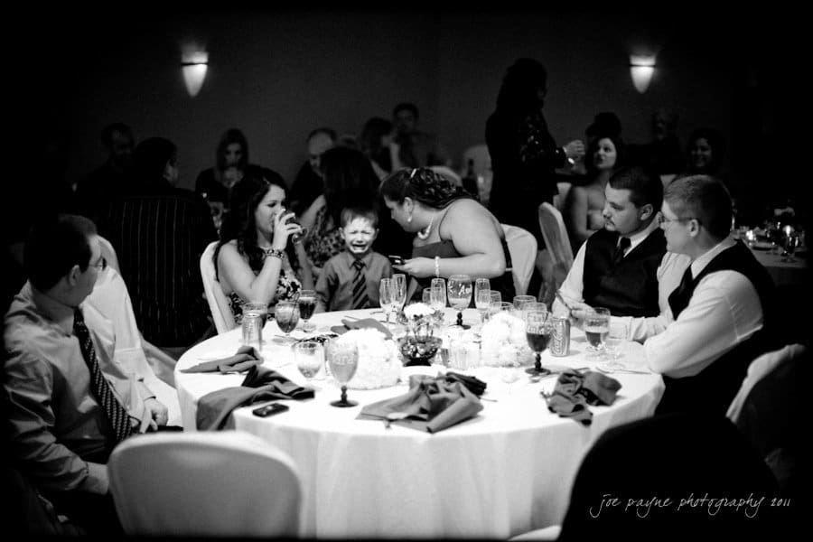 raleigh wedding photographer - jenna and ryan A black and white photo of a wedding reception. Guests are seated around a table with drinks and decorations. A young boy in a suit smiles widely, drawing attention. The atmosphere is lively and festive.