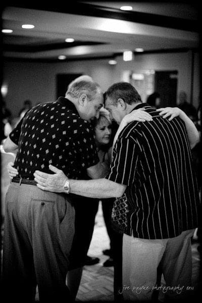 raleigh wedding photographer - jenna and ryan Three people stand in a tight circle with their arms around each other, appearing engaged in an intimate conversation at an indoor gathering. The image is in black and white, adding a timeless feel to the scene.