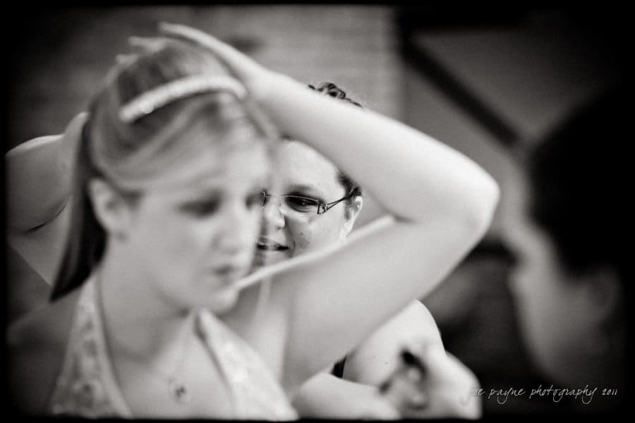 raleigh wedding photographer - jenna and ryan A black and white photo shows two women getting ready, with one adjusting her hair. The background is softly blurred, focusing on their expressions and movements.