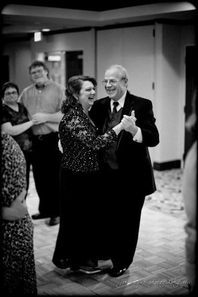 raleigh wedding photographer - jenna and ryan An elderly couple is sharing a joyful dance together at an event. The man is in a suit, and the woman is wearing a sequined dress. They are smiling, surrounded by other guests, in a warmly lit room with a patterned floor.