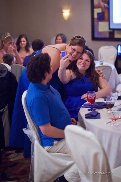 raleigh wedding photographer - jenna and ryan Two women in blue dresses take a selfie at a table during an event. A man in a blue shirt sits nearby, looking on. The room is decorated with white chair covers and a colorful abstract painting is visible in the background.