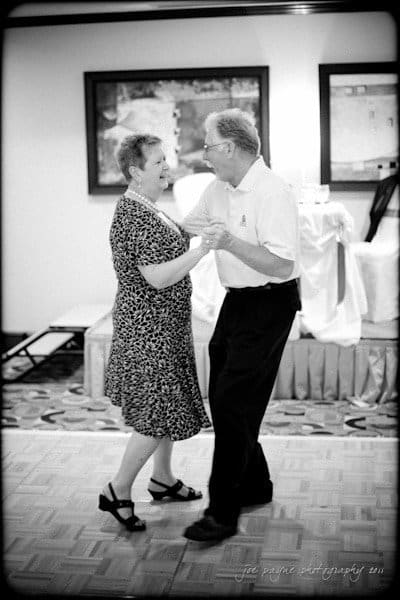 raleigh wedding photographer - jenna and ryan An elderly couple joyfully dances together on a wooden floor in a warmly lit room. The woman wears a patterned dress and the man is in a white shirt and dark pants. They appear to be laughing and enjoying the moment.