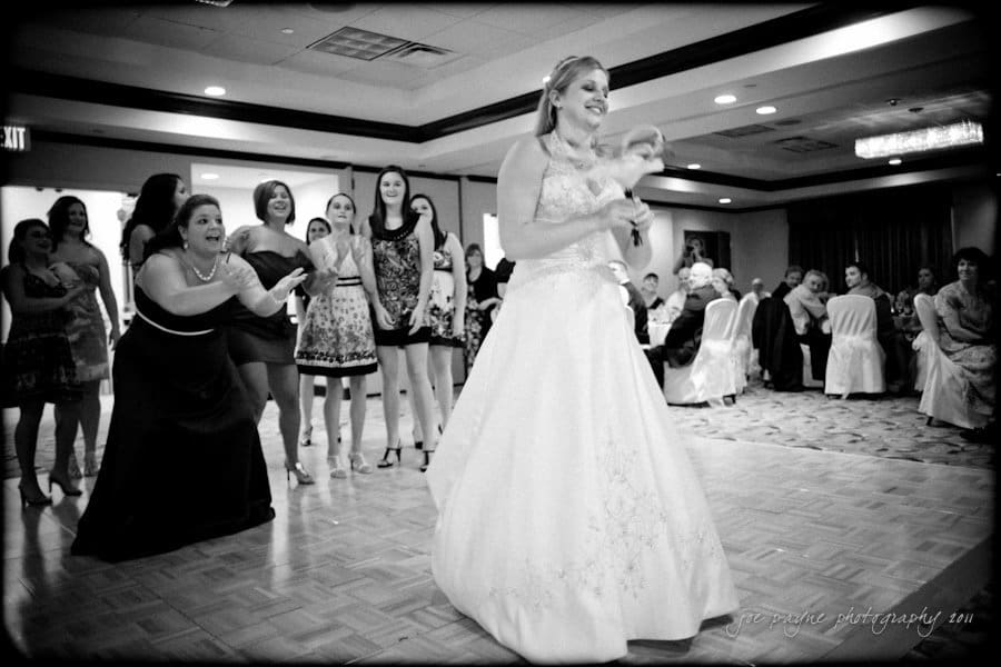 raleigh wedding photographer - jenna and ryan A bride in a white gown stands smiling, holding a bouquet in a banquet hall. Behind her, a group of women eagerly anticipate the bouquet toss. The scene is lively, with people seated at tables in the background.