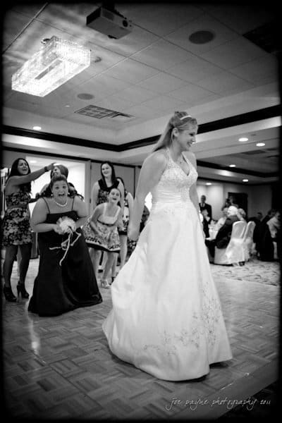 raleigh wedding photographer - jenna and ryan A black and white photo of a bride preparing to toss her bouquet at a wedding reception. Several women stand behind her, eagerly anticipating the moment. The setting is a spacious event hall with overhead lighting.