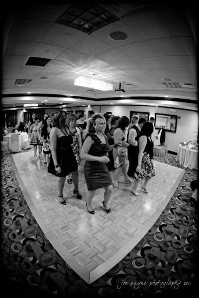 raleigh wedding photographer - jenna and ryan A group of people, dressed in formal attire, are dancing in formation on a parquet dance floor inside a well-lit room. The photo is black and white and has a fisheye lens effect, capturing a diverse group of individuals enjoying the moment.
