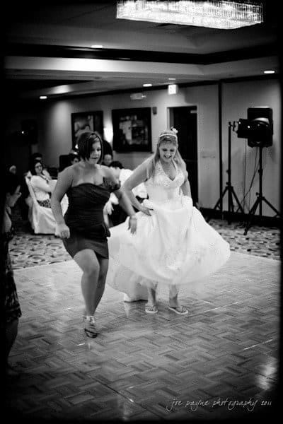 raleigh wedding photographer - jenna and ryan A bride in a wedding gown and a woman in a short dress dance energetically on a wooden dance floor at a wedding reception. Guests are seated in the background, and the room is elegantly decorated.