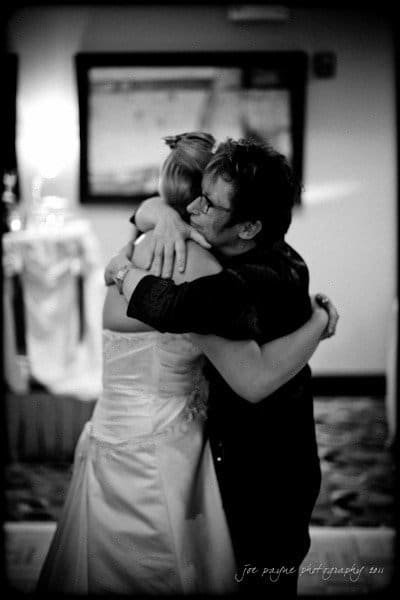 raleigh wedding photographer - jenna and ryan A couple shares a heartfelt hug in a warmly lit room. The woman is wearing a wedding dress, and the man is in dark attire. A mirror is visible in the background. The image is in black and white.