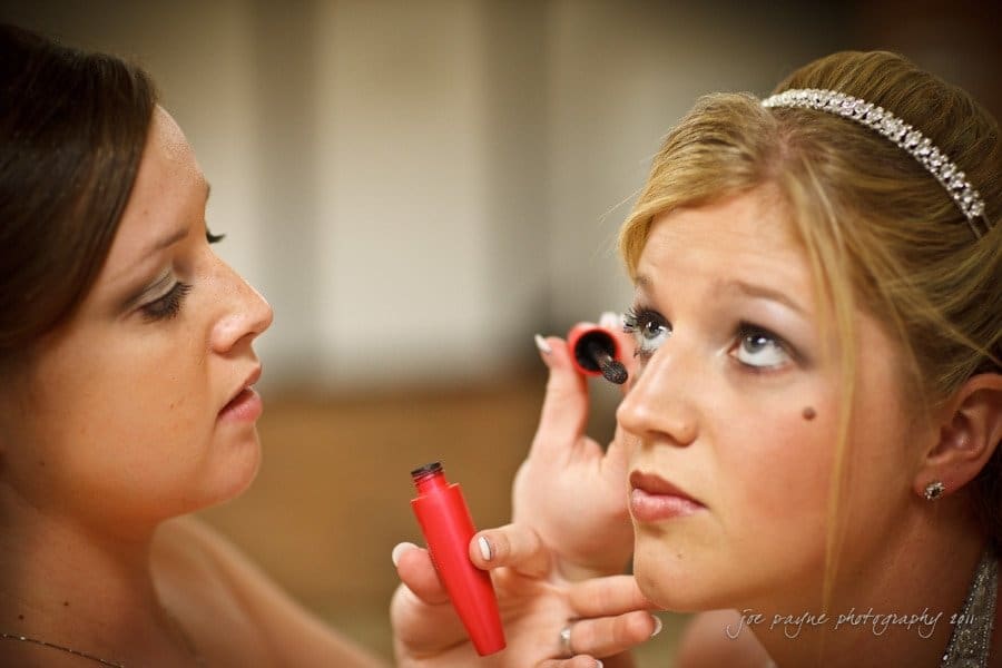raleigh wedding photographer - jenna and ryan A bride gets her makeup done by a woman applying mascara. The bride has blonde hair styled elegantly with a jeweled headband, and the makeup artist is concentrating on her work. Both are in focus against a softly blurred background.