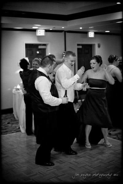 raleigh wedding photographer - jenna and ryan A black and white photo of three people dancing at a party or wedding. One person is holding a bottle, and they all appear joyful. In the background, more people are gathered, and tables are visible.