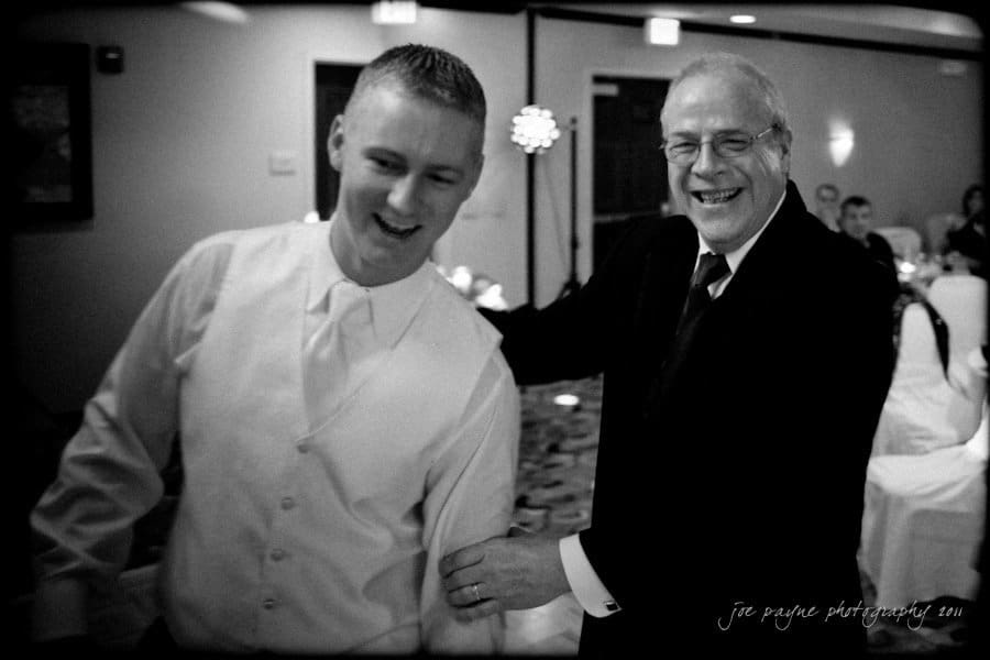 raleigh wedding photographer - jenna and ryan A joyful moment captured at a formal event, showing two men dressed in suits. They are laughing and engaged in a lively interaction, with tables and chairs in the background creating a warm, celebratory atmosphere.