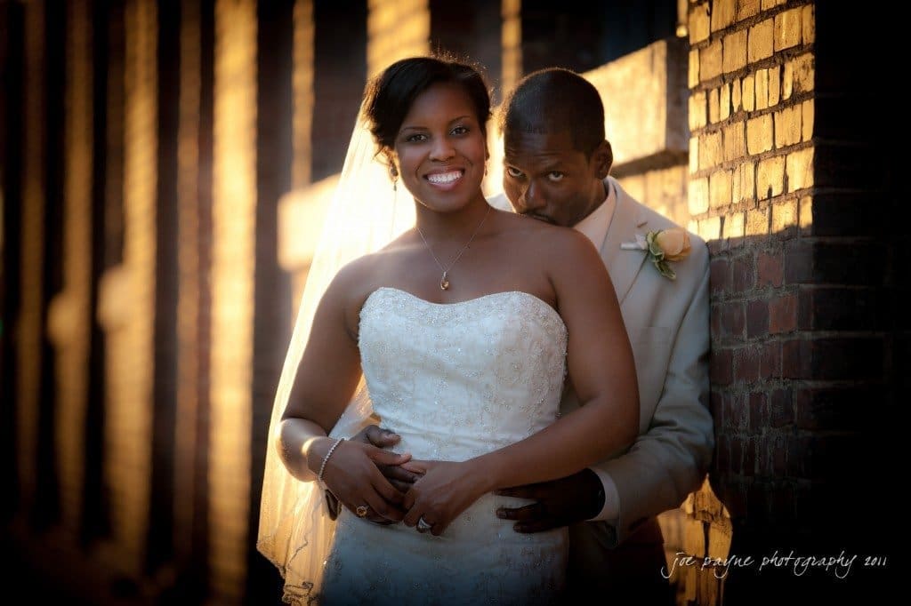 raleigh wedding photographer - image of the week: no. 5 A bride and groom pose outside against a brick wall. The groom stands behind the bride, both smiling warmly. The bride wears a white strapless gown and veil, while the groom is in a light-colored suit with a rose boutonniere.