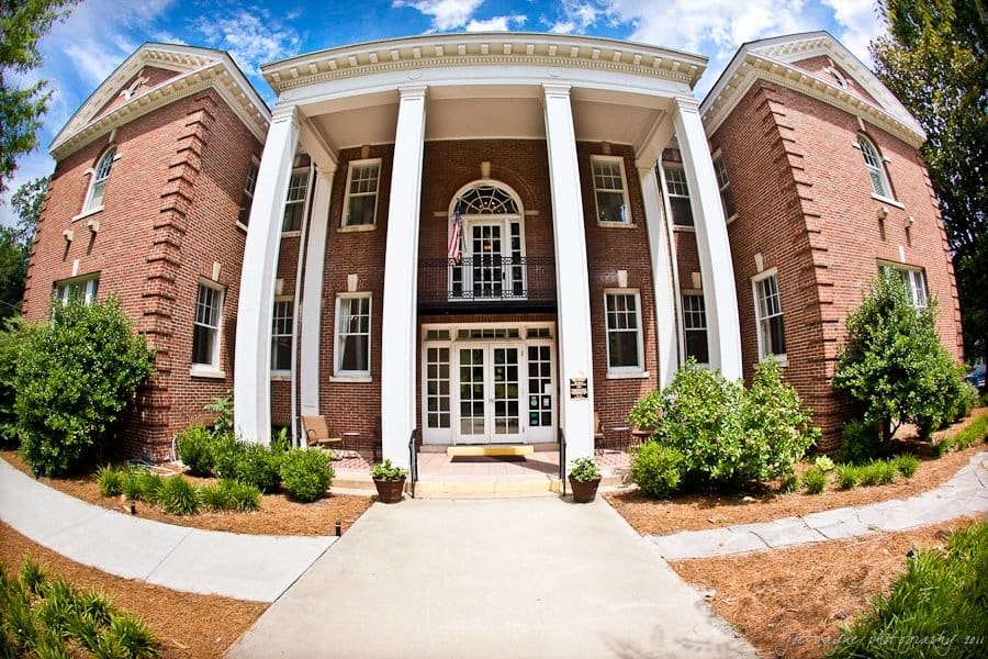 A wide-angle view of a red brick building with tall white columns at the entrance. The facade features multiple windows, and there are small bushes lining the walkway leading to the door. The sky is partly cloudy, providing a bright backdrop.