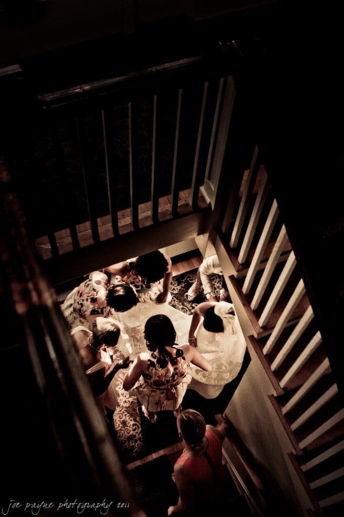 A bride in a white dress is surrounded by bridesmaids on a staircase, as seen from above. The image has a warm, vintage ambiance.