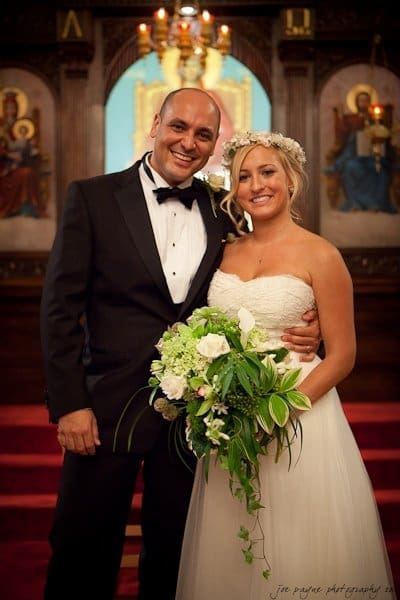A couple stands side by side, smiling at their wedding. The groom is in a black tuxedo, and the bride is in a white dress with a floral crown, holding a bouquet. They are in a church with an ornate background.