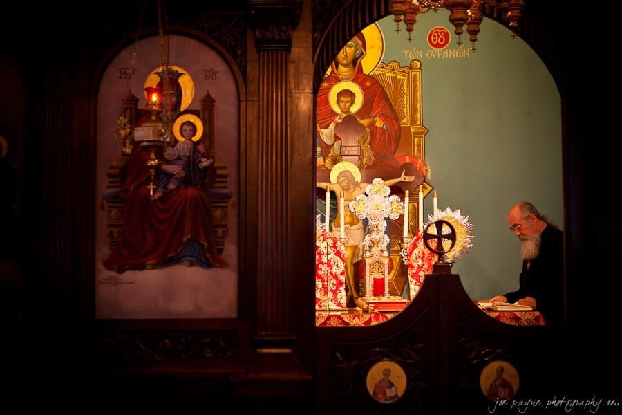 A man stands in a dimly lit orthodox church, surrounded by religious icons and ornate decorations. On the left, an icon of a seated saint with a halo is visible. The atmosphere is solemn and contemplative.