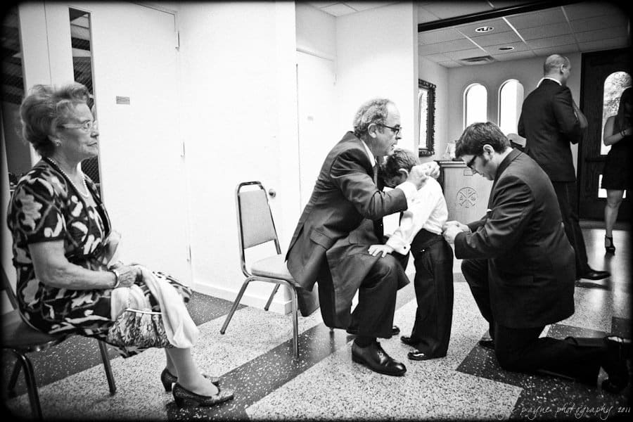 A black-and-white photo shows two men kneeling, blessing a child who is kneeling with them. An elderly woman sits on a chair watching the scene. In the background, a few people stand and converse in a room with arched windows.