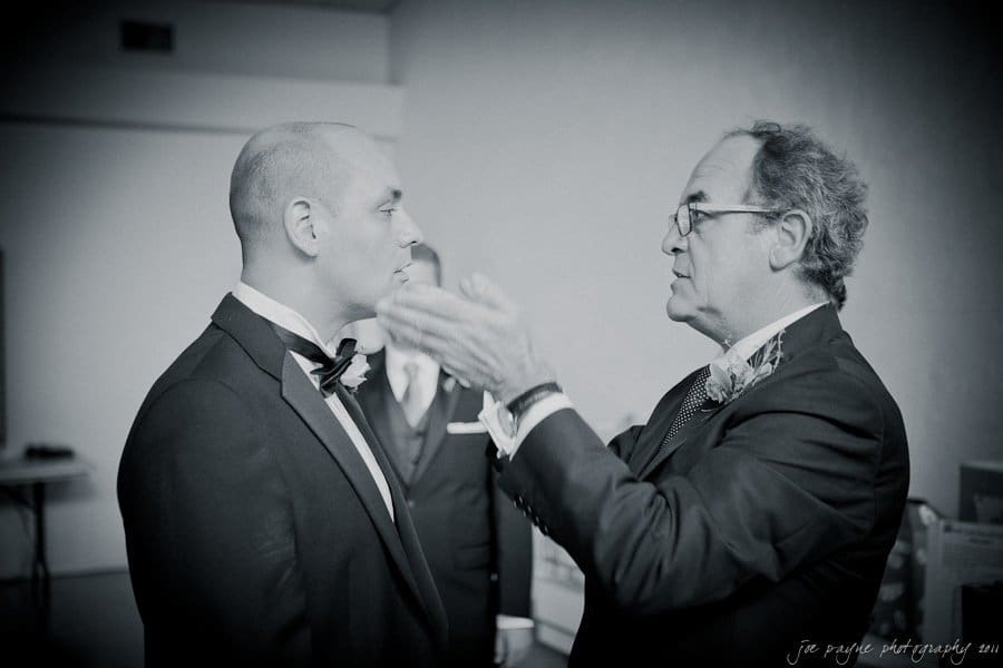 A man in a suit and bow tie receives encouragement from an older man in glasses and a suit. The elder has his hands up as if offering advice. They are indoors with a third person partially visible in the background. The photo is in black and white.