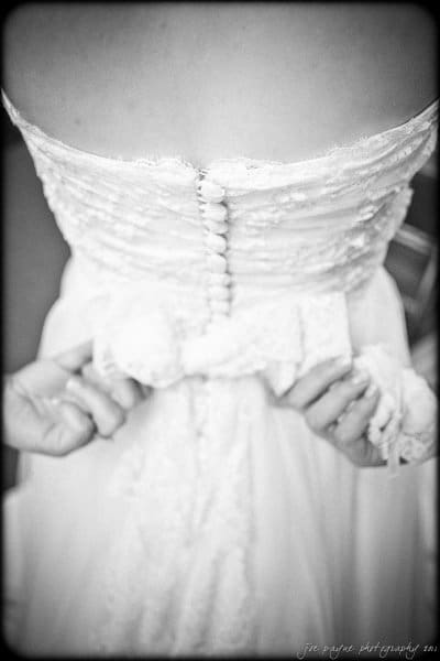 Black and white image of a bride from the back. She wears a strapless wedding dress with button details. She is adjusting her dress, holding its fabric with both hands.