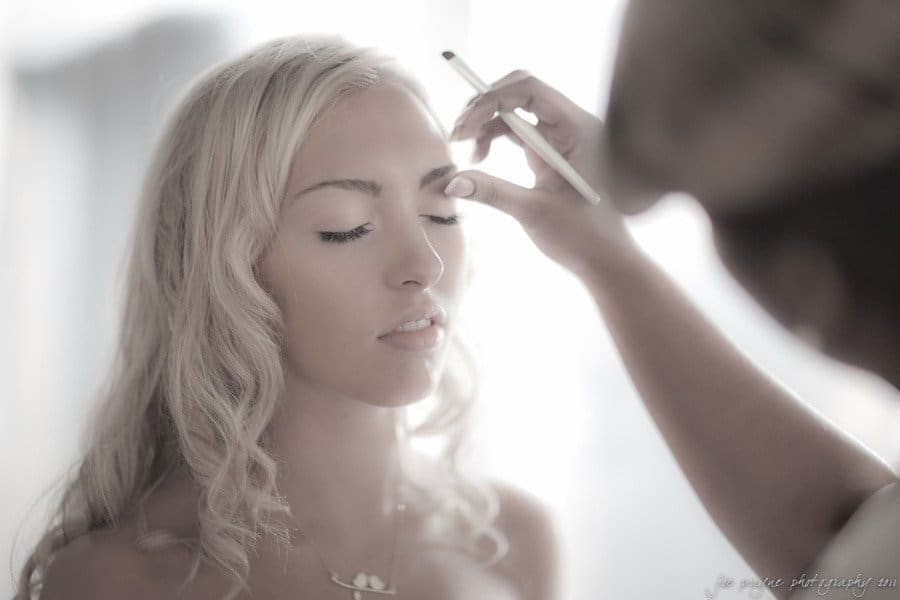 A woman with long blonde hair is having her makeup applied with a brush. She has her eyes closed and appears relaxed. The background is softly blurred, giving the image a serene, ethereal quality.