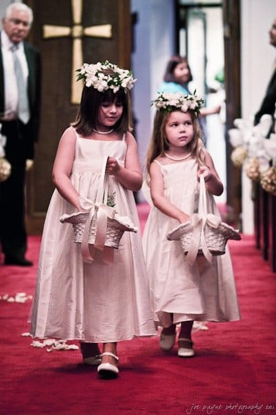 Two young flower girls dressed in white dresses walk down a red-carpeted aisle carrying white baskets. They wear flower crowns and look focused. A cross and a few guests are in the background.