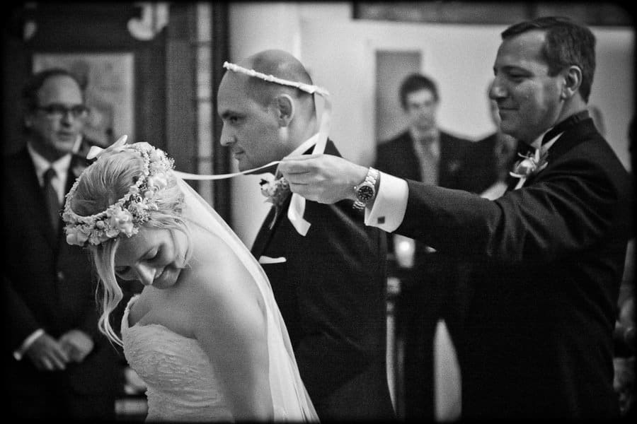 A black and white photo of a wedding ceremony where a man places a crown connected by a ribbon on the heads of a bride and groom. The bride smiles while the groom stands beside her. Onlookers are visible in the background.