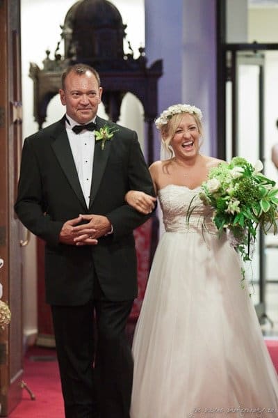 A bride in a white dress and flower crown walks arm in arm with a man in a black suit and bow tie. They are smiling and appear to be in a church setting. The bride holds a bouquet of green and white flowers.