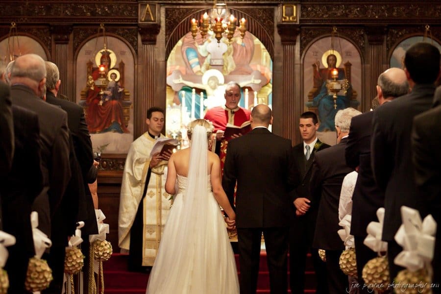 A couple stands at the altar of a decorated church, facing a priest in red robes. Guests, dressed in formal attire, are seated on both sides of the aisle. Liturgical icons adorn the background.