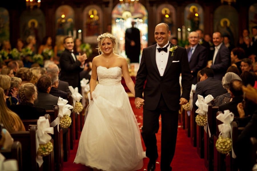 A bride and groom joyfully walk down the aisle in a church. The bride wears a white gown and floral headpiece, while the groom is in a black tuxedo. The pews are filled with seated guests, decorated with white bows and flowers.