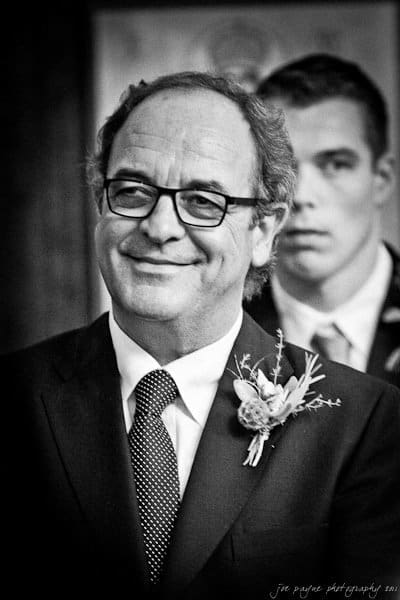 A man wearing glasses and a suit with a boutonniere smiles warmly. In the background, another person in a suit looks on. The image is black and white, creating a classic and timeless feel.