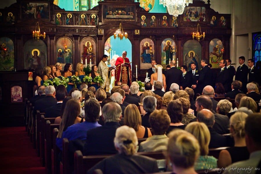 A wedding ceremony in a church, with the bride, groom, and wedding party standing at the altar. Guests are seated, watching the proceedings. The church is adorned with religious icons and a chandelier.