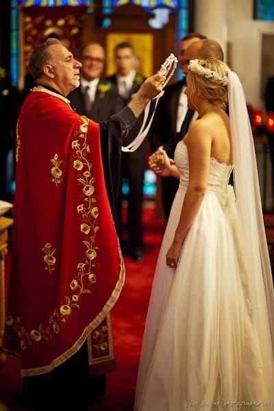 A bride and groom stand in a church during a wedding ceremony. A priest in a red robe holds a cross and chain above them. The bride wears a white gown and veil. Stained glass windows are visible in the background.