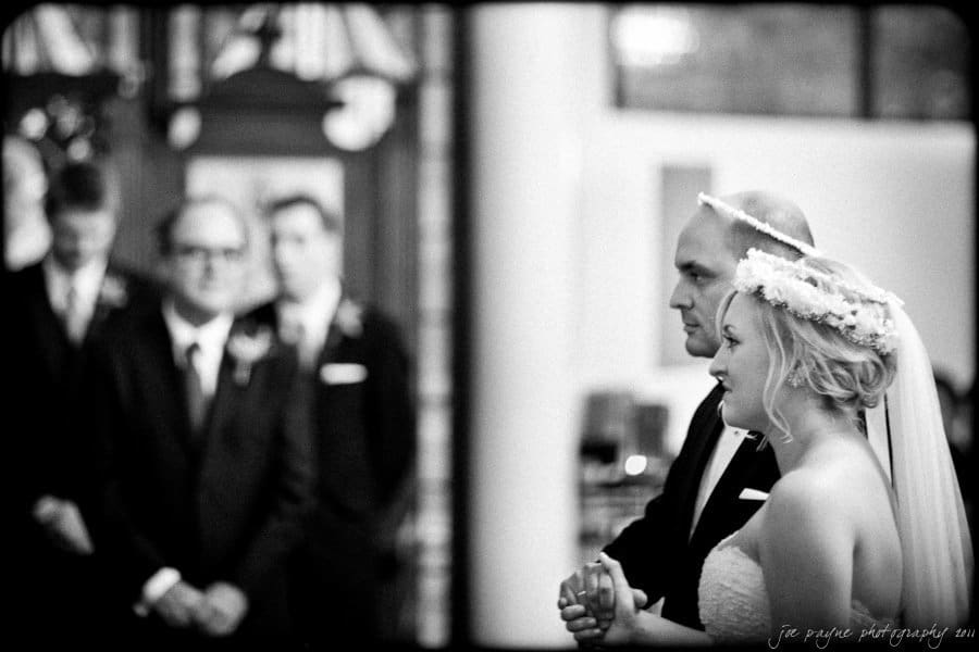 A bride and groom stand in a church, holding hands during their wedding ceremony. She wears a floral crown and veil, while he is in a suit. The background is softly blurred, showing guests attentively watching.