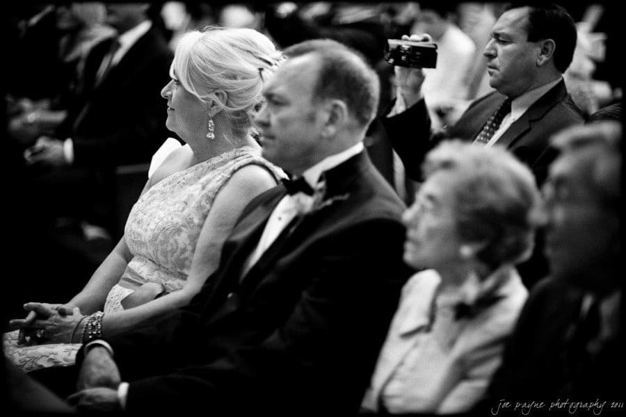 A black and white photo of several people seated in an audience. In the foreground, two elegantly dressed individuals, a woman and a man, are watching intently. Behind them, a man is taking a picture with a camera.