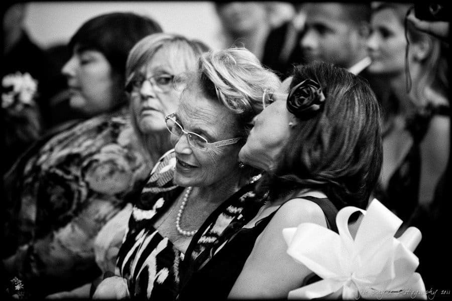 A black and white photo of two women sitting closely among a crowd. One woman whispers into the others ear. The older woman wears glasses and a patterned dress. The woman whispering has a large bow on her shoulder. Other attendees are visible in the background.