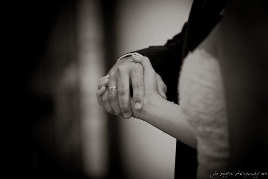A black and white photo of two people holding hands. One is wearing a suit with a visible ring, and the other is in a textured, white dress. The focus is on their intertwined hands, suggesting connection or a moment of intimacy.