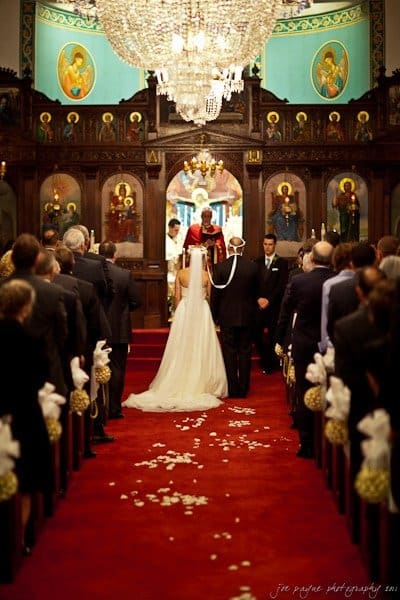 A bride and groom stand before a priest in a richly decorated church, under an ornate chandelier. Guests fill the pews along a red carpet aisle adorned with white bows and rose petals. Sacred artwork and icons are visible on the walls.