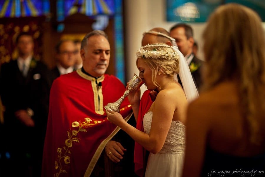 A bride in a wedding gown and veil drinks from a silver chalice during a ceremony. A man in a red ornate robe stands beside her. Onlookers are in the background with stained glass windows visible.
