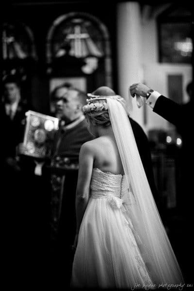 A bride in a strapless gown stands indoors with her back to the camera. She wears a long veil and floral headpiece. Another person holds an object above her head during a ceremony. The setting is formal with blurred figures in the background.