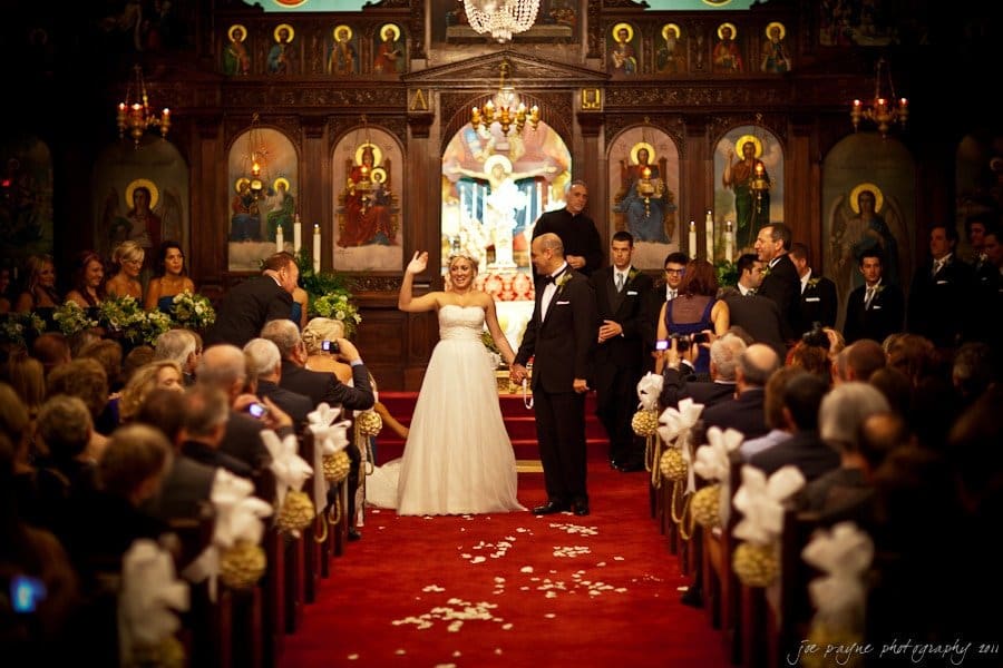 A bride and groom stand at the altar in an ornate church, surrounded by their wedding party and seated guests. The aisle is decorated with white bows, and the church is adorned with colorful religious icons.