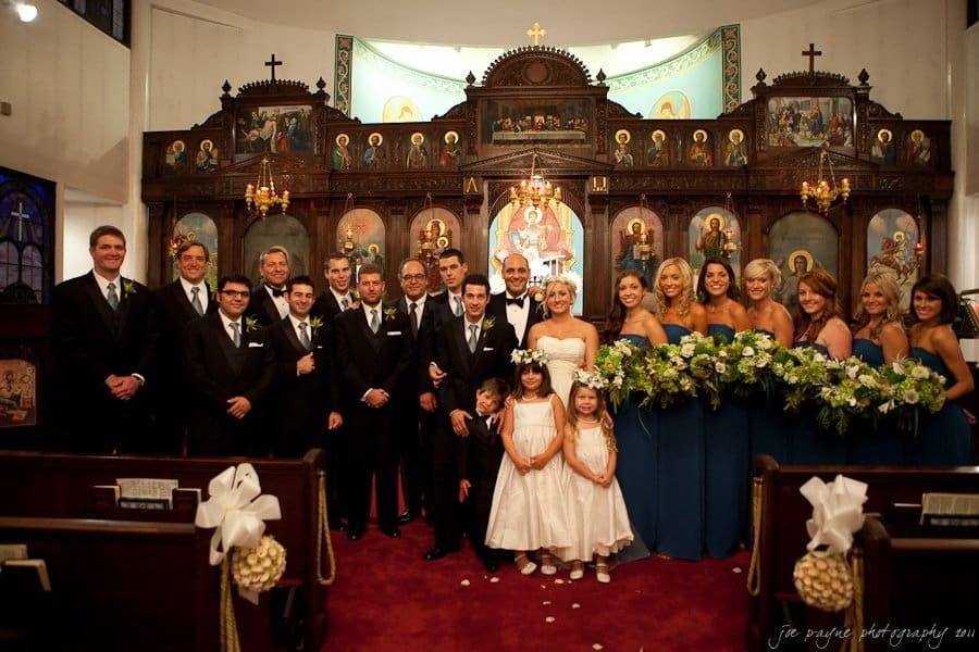 A wedding party posed inside a church with ornate wooden decorations and religious icons. The group includes bridesmaids in blue dresses, groomsmen in black suits, two flower girls in white dresses, and the bride and groom at the center.