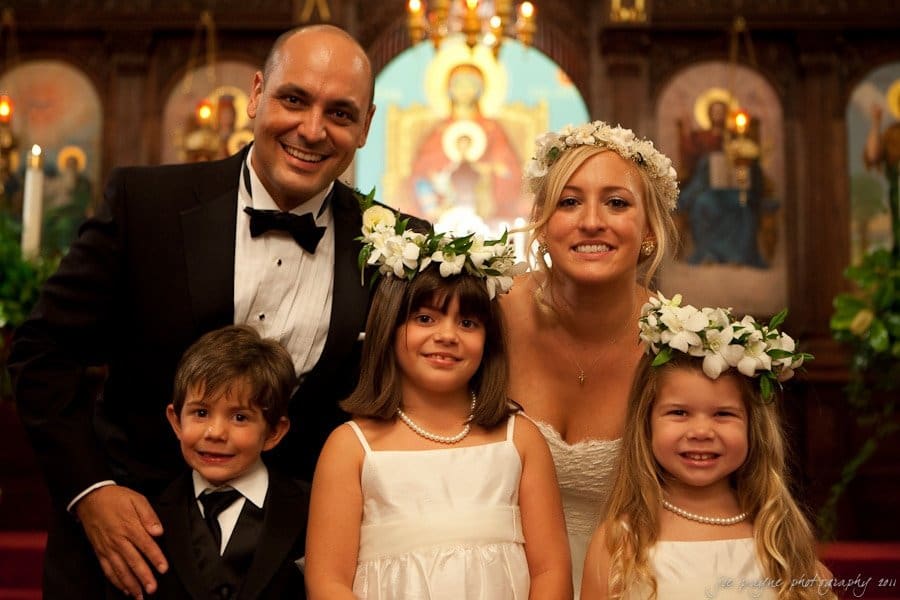 A bride and groom smile with three children at a wedding ceremony. The bride wears a floral crown and a white dress, while the groom is in a black tuxedo. The children, two girls and a boy, wear white outfits and flower crowns. An altar is in the background.