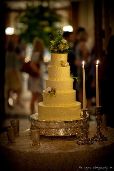 A five-tier yellow cake decorated with flowers stands on a silver platter. Two lit candles in ornate holders sit beside it, creating a warm glow. The background is softly blurred with people and greenery.