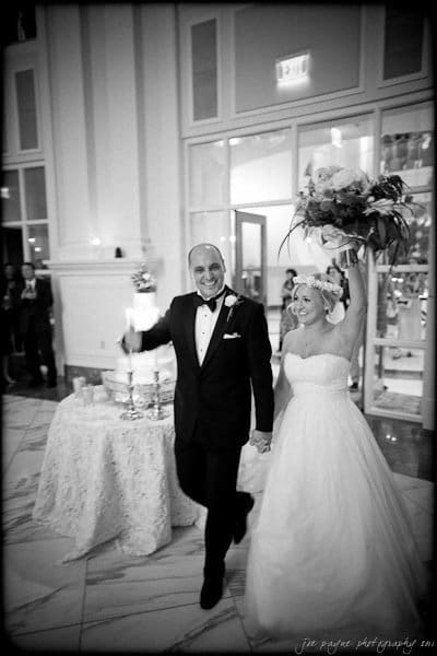 A bride and groom smile and hold hands as they enter a room. The bride, wearing a strapless gown, extends her arm holding a bouquet. The groom is in a tuxedo. A decorated cake is visible on a table in the elegant setting.