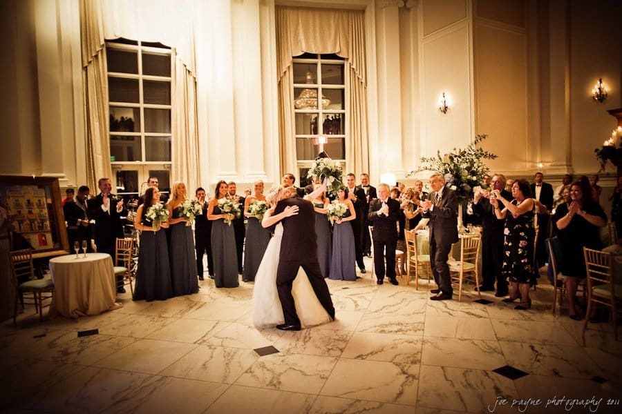 A bride and groom share their first dance in an elegant ballroom, surrounded by their wedding party and guests. The room features high ceilings, large windows, and chandeliers. Bridesmaids in dark dresses and groomsmen in suits look on and applaud.