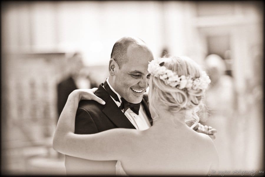 A couple sharing a dance in a sepia-toned image. The man is smiling warmly, wearing a tuxedo, while the woman, with blonde hair and a floral crown, faces him. The background is softly blurred, highlighting their connection.