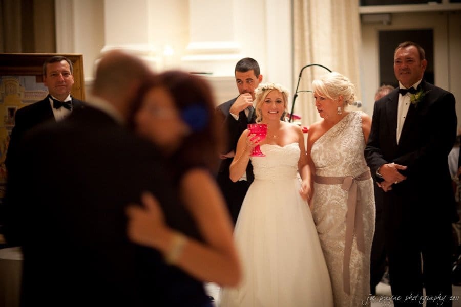 A bride in a white gown smiles while holding a pink drink, surrounded by two women and two men in formal attire. In the foreground, a couple dances. The setting appears to be an elegant indoor venue.