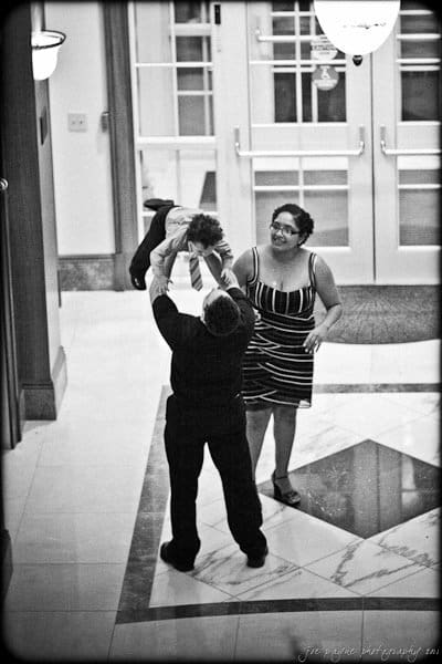A man playfully lifts a child in the air while a woman smiles nearby. They stand in a hallway with marble flooring and glass doors in the background. The image is in black and white.