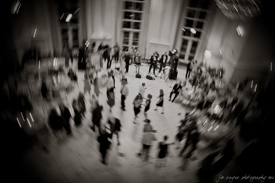 A black and white image of a large, elegant ballroom with a blurred motion effect. Many people are dancing and mingling in formal attire. A band is performing in the background near tall windows.